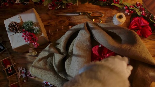 A top view of Santa's gloved hands folding gifts into his sack on a wooden table among cards, paints, and garlands, symbolizing the arrival of Christmas and preparations for the holiday.