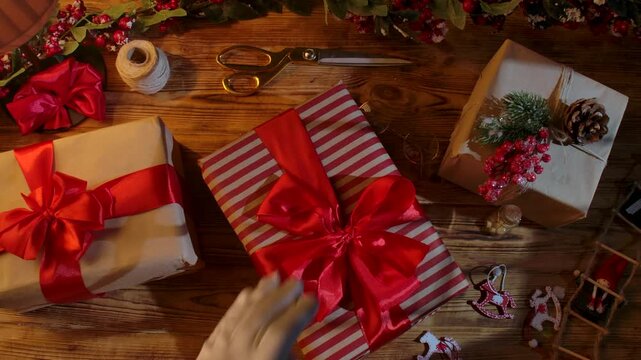 Top view of Santa Claus's gloved hands arranging festive gift boxes on a rustic wooden table. Preparations for New Year's and Christmas celebrations.