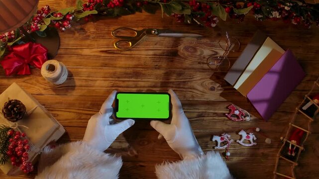 Top view of Santa Claus's gloved hands holding a smartphone with a green chroma key screen on a rustic wooden table. Preparing for New Year's and Christmas celebrations.