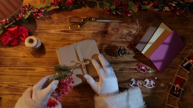 Top view of Santa Claus's gloved hands creating festive gifts with his own hands on a rustic wooden table. Preparing for New Year's and Christmas celebrations.