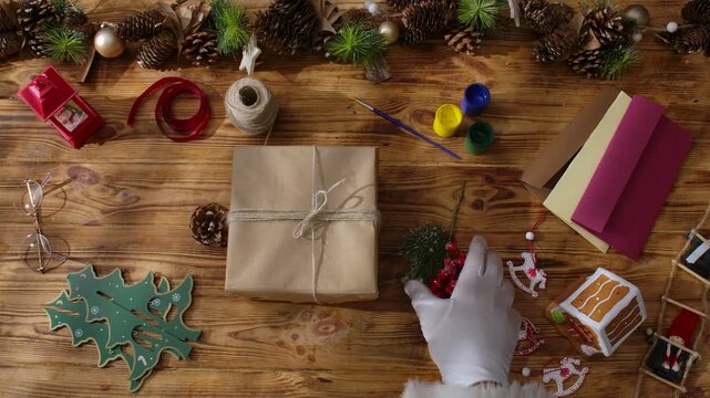 Top view of Santa Claus's gloved hands creating festive gifts with his own hands on a rustic wooden table. Preparing for New Year's and Christmas celebrations.