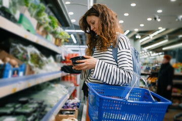 Woman carefully inspecting a pre-packaged food product in the fresh produce section. Focusing on making an informed purchase decision while grocery shopping in a brightly lit modern supermarket aisle