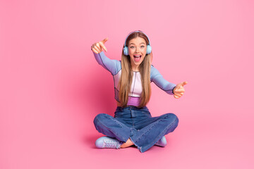 Adorable blonde girl with headphones sitting cross-legged on a pink background, expressing joy and...