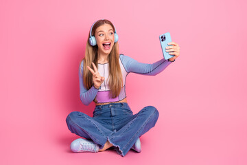 Charming young girl taking a joyful selfie with a smartphone while seated against a vibrant pink...