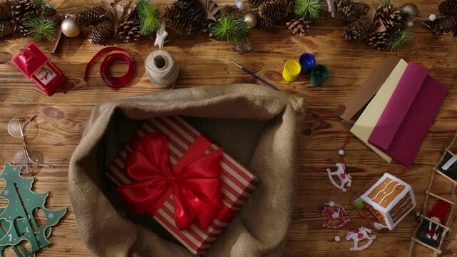 A top view of Santa's gloved hands folding gifts into his sack on a wooden table among cards, paints, and garlands, symbolizing the arrival of Christmas and preparations for the holiday.