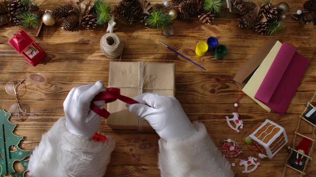 Top view of Santa Claus's gloved hands creating festive gifts with his own hands on a rustic wooden table. Preparing for New Year's and Christmas celebrations.