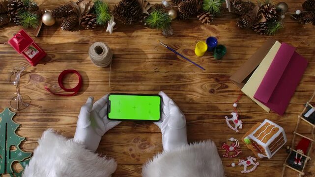 Top view of Santa Claus's gloved hands holding a smartphone with a green chroma key screen on a rustic wooden table. Preparing for New Year's and Christmas celebrations.