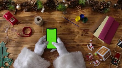 Top view of Santa Claus's gloved hands typing on a smartphone with a green chroma key screen on a rustic wooden table. Preparing for New Year's and Christmas celebrations. - Powered by Adobe