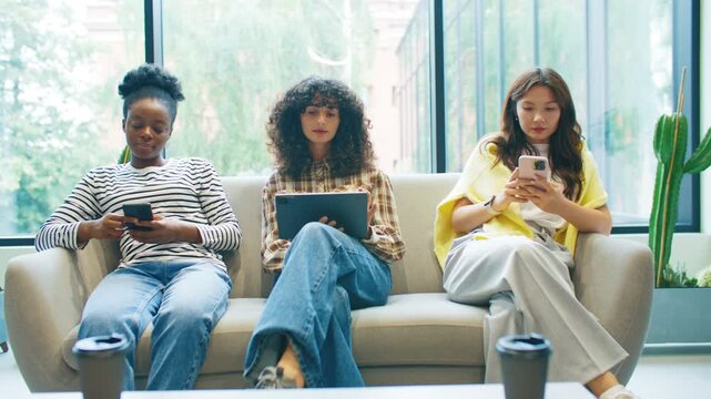 Three women sitting on sofa in bright waiting room using digital devices. Female holding tablet reading while others scrolling smartphones. Modern lounge with daylight and calm atmosphere.
