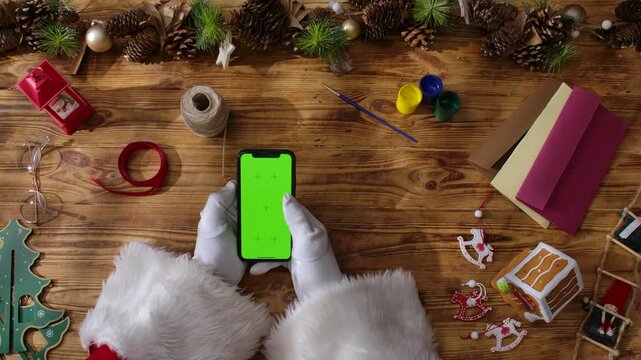 Top view of Santa Claus's gloved hands swiping on a smartphone with a green chroma key screen on a rustic wooden table. Preparing for New Year's and Christmas celebrations.