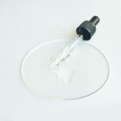 Transparent dropper with cosmetic serum on clear petri dish on white background.