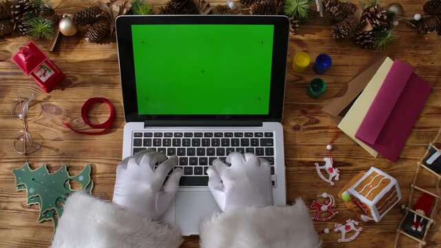 Top view of Santa's gloved hands typing on a laptop keyboard with a green chroma key screen on a rustic wooden table. Preparing for New Year's and Christmas celebrations.