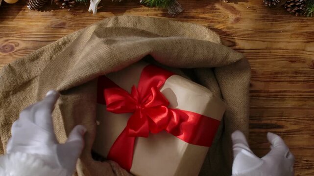 A top view of Santa's gloved hands folding gifts into his sack on a wooden table among cards, paints, and garlands, symbolizing the arrival of Christmas and preparations for the holiday.