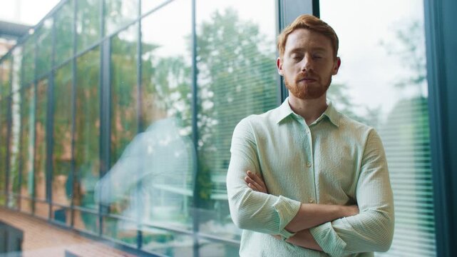 Serious man standing with folded arms near glass wall in modern office. Confident professional looking at camera showing calm focus and leadership. Natural daylight reflections on windows.