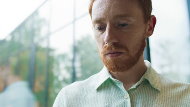 Close facial portrait of man in bright modern office near glass wall. Male getting distracted from tablet and looking directly at camera. Serious expression under soft natural daylight reflections.