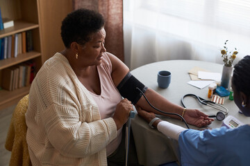 Middle aged Black woman sitting at table having blood pressure measured by female healthcare worker, holding cane in left hand, medical equipment and medication visible on table