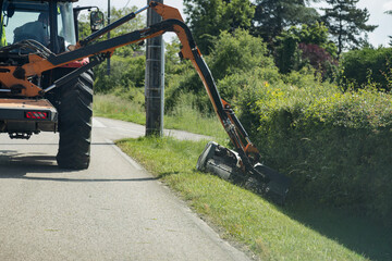 A tractor with an orange boom arm operates a hedge trimmer or flail mower, clearing vegetation from a ditch alongside an asphalt road. © DBA