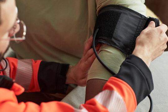 Caucasian young male paramedic wearing glasses measuring blood pressure of Black middle aged woman patient using digital monitor, focusing on arm and medical procedure in indoor setting