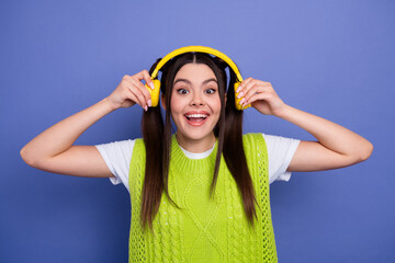 Bright cheerful young woman with yellow headphones wearing lime knit vest smiles at camera in a blue studio background