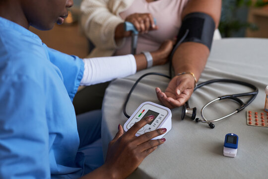Black female nurse measuring blood pressure of geriatric female patient using digital monitor, stethoscope and pulse oximeter on table, arm extended for examination