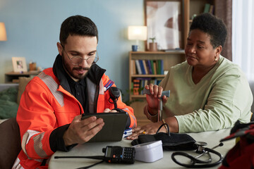 Young Caucasian male paramedic showing digital tablet to middle aged Black woman using crutch, both...