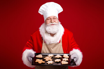 Santa in red suit and chef hat offers a tray of cookies against a deep red background perfect for Christmas market family