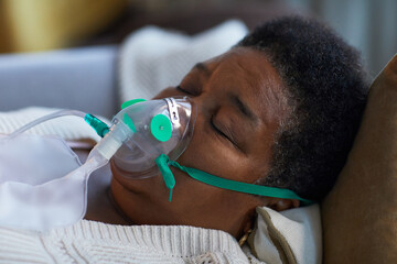 Senior Black woman lying on couch wearing oxygen mask, eyes closed, resting during illness or recovery, close up showing facial features and medical equipment, home environment
