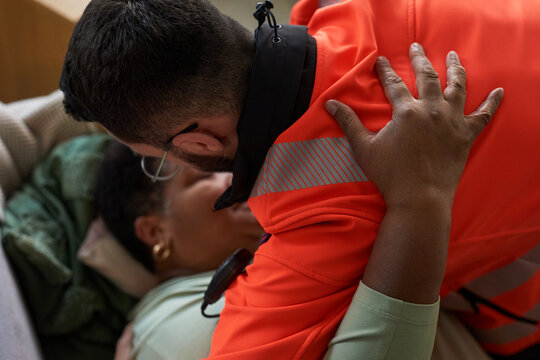 Middle aged Black woman lying on couch being assisted by young adult Caucasian male paramedic wearing uniform, paramedic leaning over providing emergency medical care