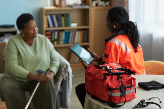 Black female paramedic showing digital tablet to senior woman with disability with cane during home visit, red medical bag and stethoscope on table in foreground, bookshelf in background
