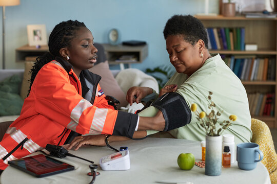 Black female healthcare worker measuring blood pressure of middle aged Black woman, using digital monitor, both sitting at table in home setting, medical equipment visible on table - Powered by Adobe