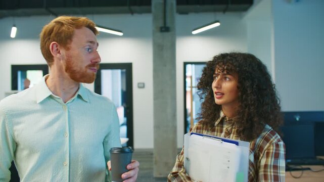 Coworkers talking while walking. Man with red hair holding coffee cup and woman with curly hair carrying documents. Talking seriously. Discussing work matters together inside office workspace.