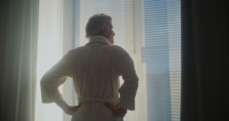 Mature Man Looks Out Window, Pulls Back Heavy Curtains, and Stretches. Modern Hotel Room Filled With Bright Sunlight. Concept of Relaxation, Travel, Good Morning, Emotional Well Being. Back View.