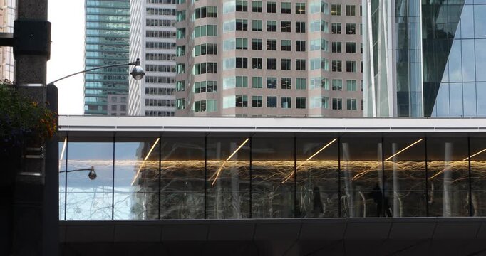 Glass pedestrian skywalk suspended between modern office towers in dense downtown core. Commuters walk past warm light trails, reflections, suggesting corporate routine, late work and urban isolation.