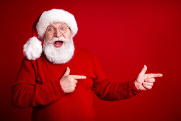 Cheerful Santa Claus points with excitement in a bright red studio setting wearing a classic red sweater and hat for festive holiday promotion