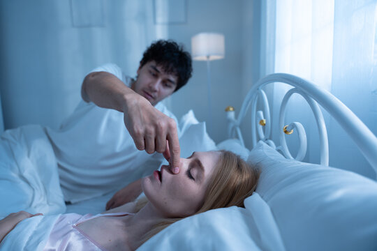 Annoyed man trying to stop his partner from snoring by pinching her nose while she sleeps, depicting sleep disturbance, relationship tension, snoring, couple, bed, sleep. woman. couple-problem.