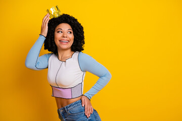 Cheerful young woman with curly hair wearing crown against a yellow background symbolizing beauty...