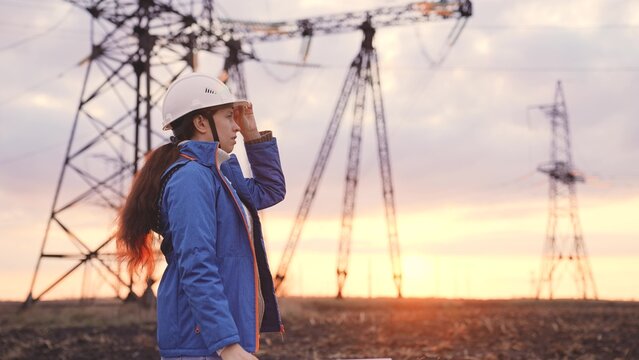 An engineer at a high-voltage power plant is working making an inspection at the sunset in the sky, electricity design, industry concept, modern business technology, an electrician with a project on - Powered by Adobe