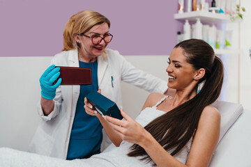 Cosmetologist in blue gloves and lab coat presenting various skincare products to a smiling female client lying on a treatment bed in a modern beauty clinic
