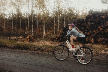 Title: A young woman on a bicycle, resting on a forest road next to a pile of cut trunks, in the warm, golden light of late afternoon, symbolizing active recreation in nature