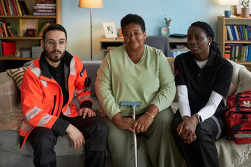 Portrait of middle aged Black woman with disability sitting on sofa holding cane, flanked by young adult Caucasian man and young adult Black woman wearing paramedic uniforms