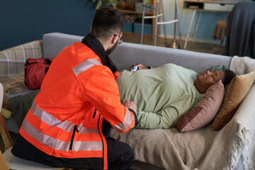 Black middle aged woman lying on couch receiving medical assistance from Caucasian middle aged male paramedic wearing high visibility jacket, paramedic checking pulse and monitoring condition