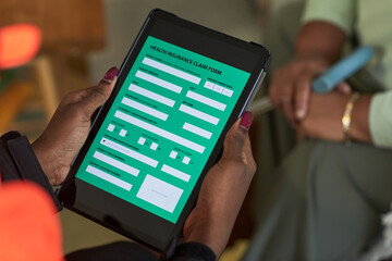 Black female nurse holding digital tablet displaying patient health insurance claim form, sitting indoors with another in background, hands visible interacting with electronic device