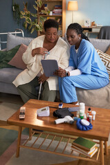Middle aged Black woman with disability sitting on sofa holding cane, consulting with young adult Black female nurse using digital tablet, medical equipment and medication on table