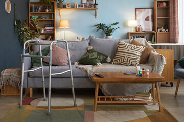 Living room scene showing walker positioned beside sofa, suggesting presence of senior with mobility challenges, coffee table holding remote control and medicine bottles, bookshelves in background