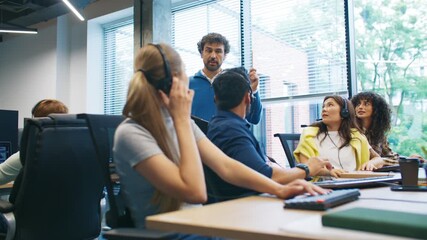 Team leader standing and assigning new tasks to call center employees wearing headsets. Customer service agents listening attentively to manager instructions during work meeting in bright office. - Powered by Adobe