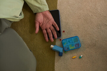 Collapsed Black woman lying on carpet with hand outstretched next to inhaler, pill organizer, scattered medication and digital tablet, suggesting medical emergency or health crisis