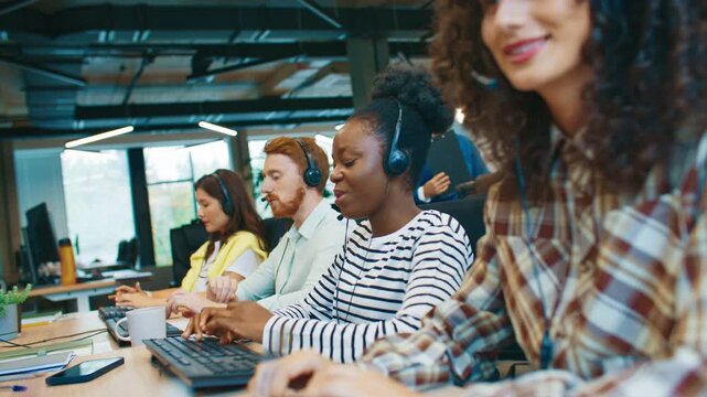 Side view of diverse call center employees wearing headsets and communicating with clients while typing. Customer support team focused on assisting callers during workday in bright open office. - Powered by Adobe