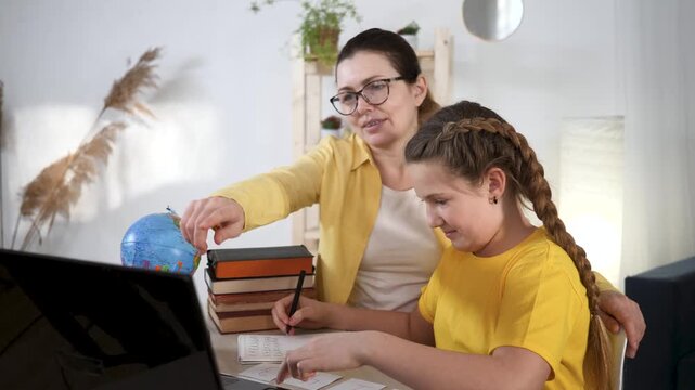 Mother tutor guides daughter studying homework at table with laptop book globe notebook and pen daughter writing notes learning focus on study for school and schoolwork with visible book and globe