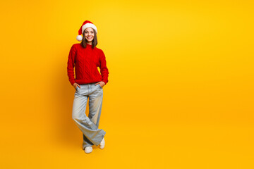 Fototapeta premium Cheerful young woman in a red Christmas sweater and Santa hat posing casually against a bright yellow background for holiday fashion and lifestyle use