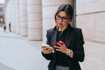 A professional woman in glasses multitasks deftly, balancing two smartphones in an urban setting, reflecting modern business dynamism.